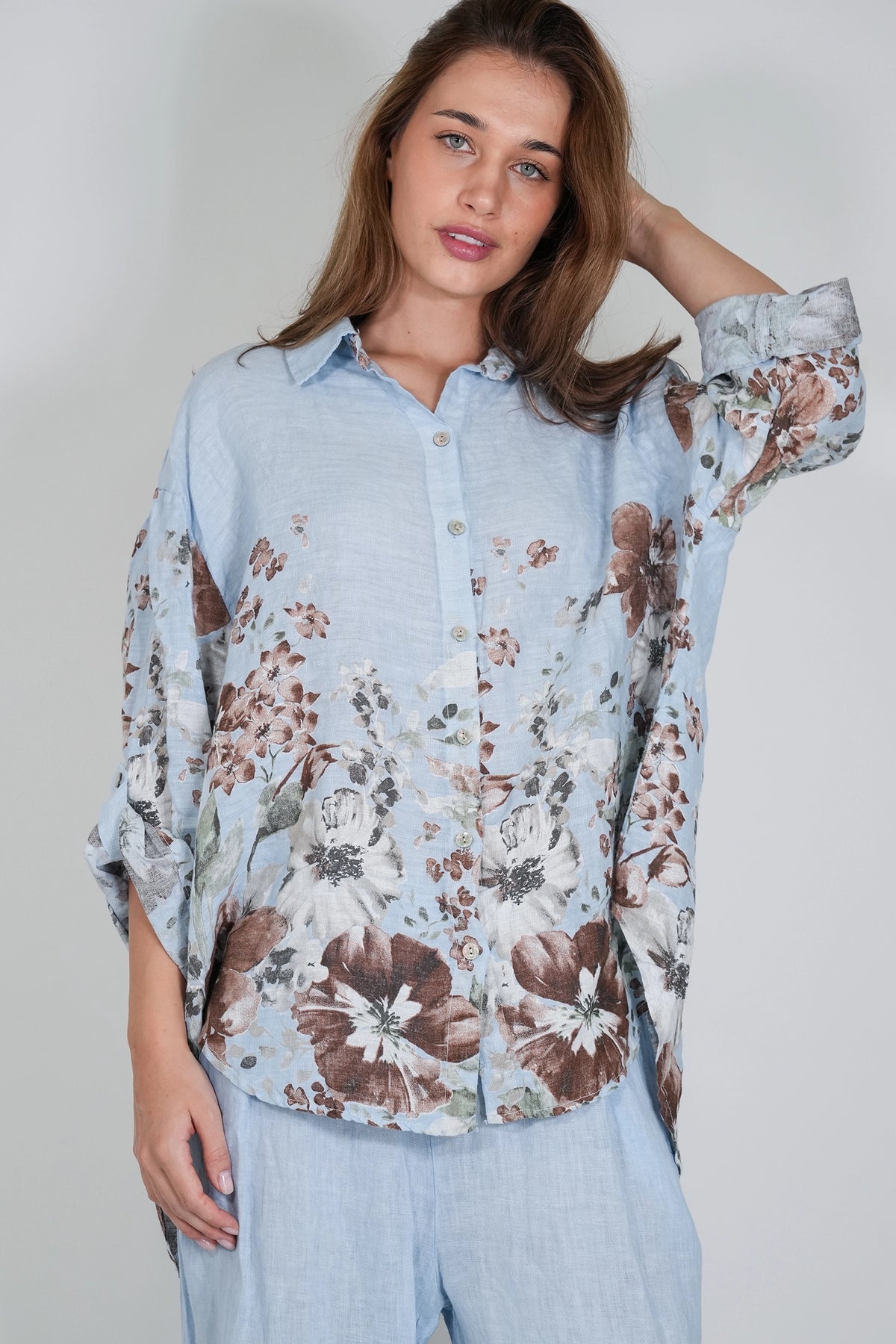 A woman wearing a light blue button-up shirt with a brown and white floral print, paired with light blue pants, against a plain light gray background.