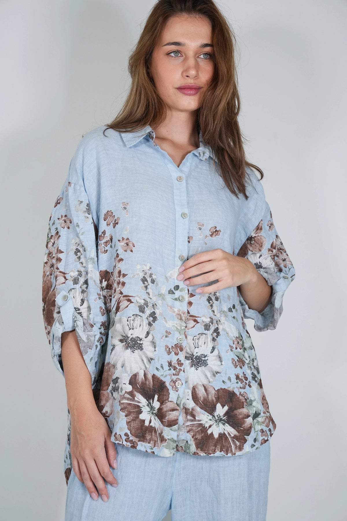 A woman wearing a light blue button-up shirt with a brown and white floral print, paired with matching light blue pants, photographed against a neutral light gray background.