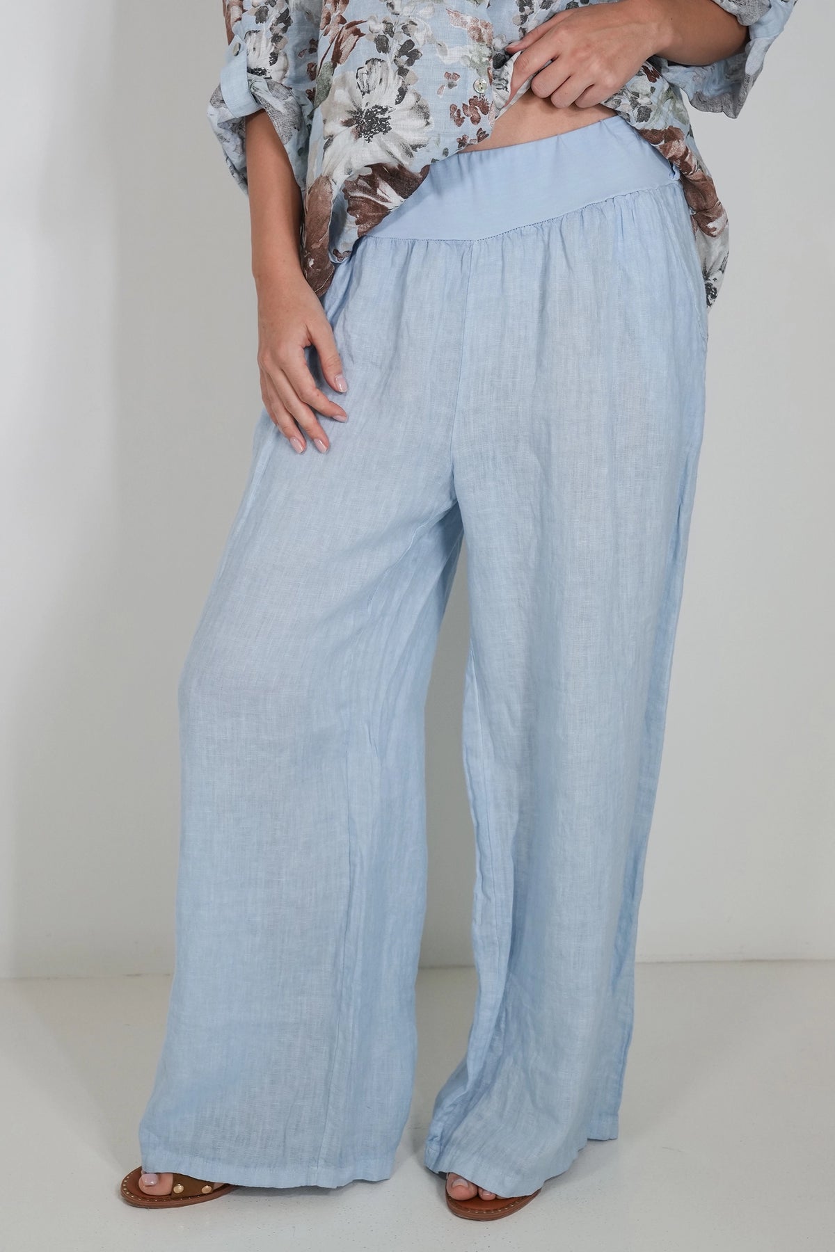 A person wearing light blue wide-leg linen pants with a matching elastic waistband, paired with a floral print top and brown sandals, shown from the waist down against a plain light background.