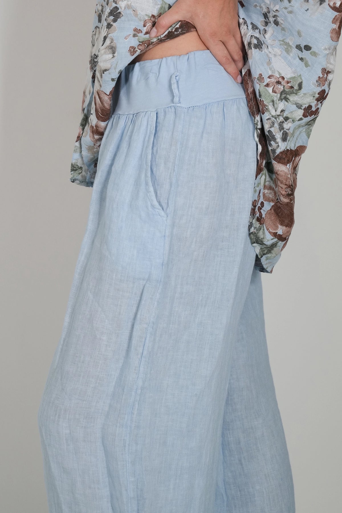 A close-up of a person wearing light blue wide-leg linen trousers, paired with a floral-patterned blouse, against a neutral light gray background.