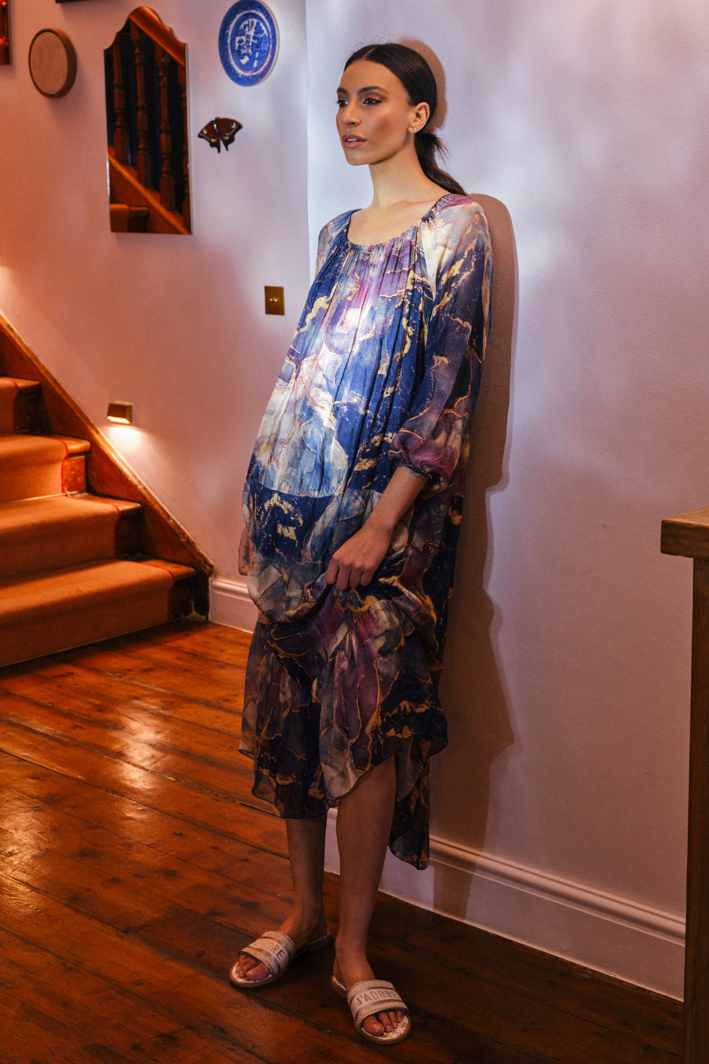 A woman stands indoors against a pale wall wearing a loose, flowy dress with an abstract painterly print in blue, purple, and white. The room has wooden stairs, a wood floor, and wall decor.