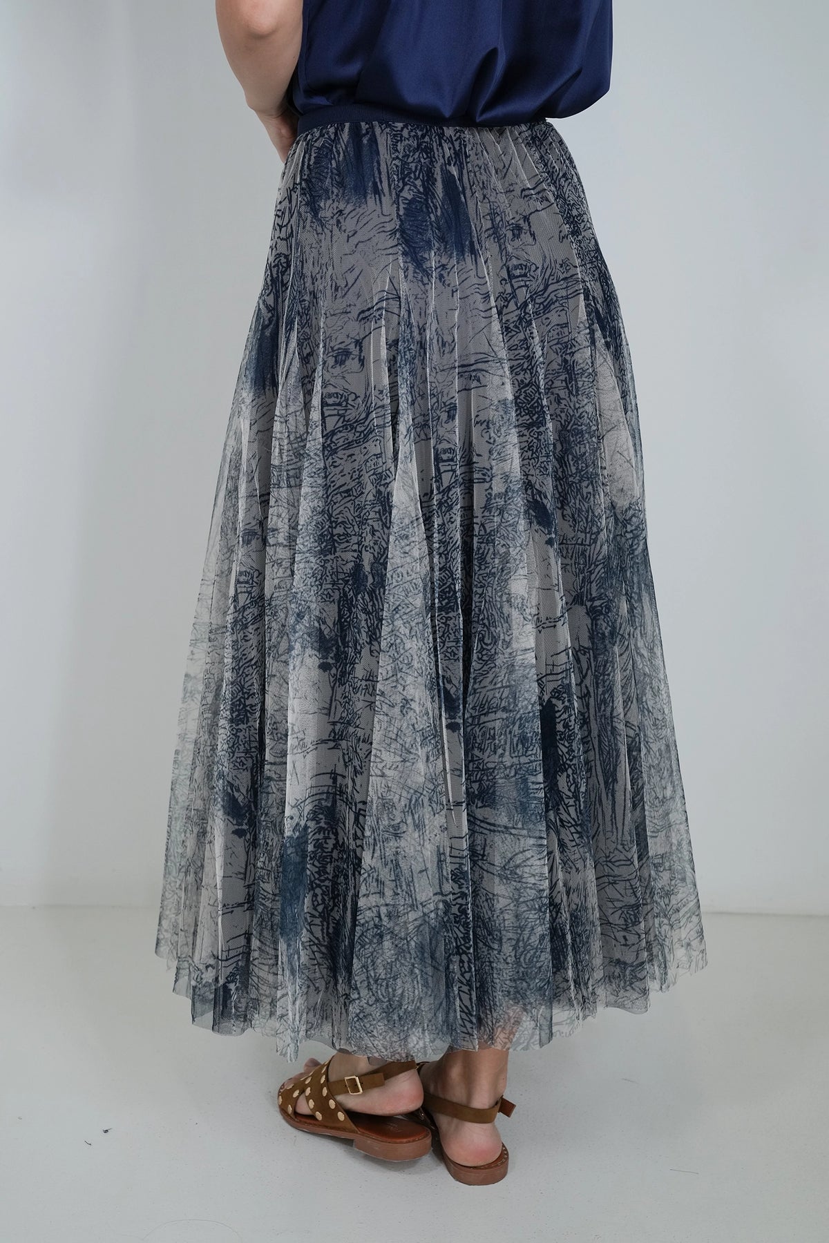 A person shown from the back wearing a long pleated tulle skirt with an abstract blue and gray print, paired with a navy top and brown sandals, against a plain light gray studio background.