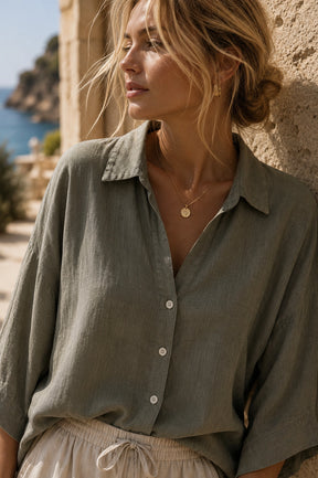 A woman wearing a loose, short-sleeve button-up shirt in a muted sage green color, leaning against a textured stone wall with a coastal background visible.