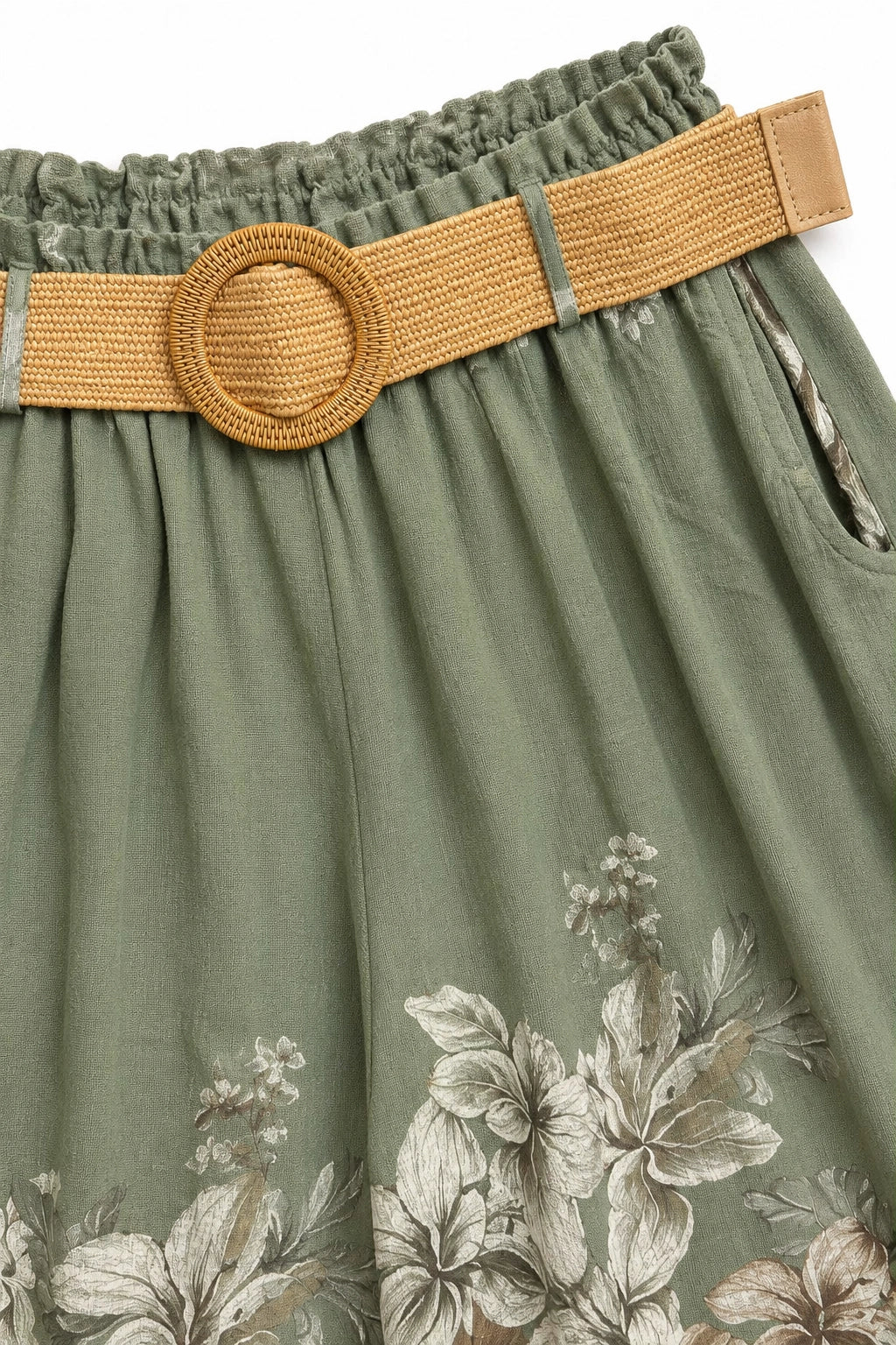 Close-up of a green floral skirt with a wide woven tan belt featuring a round woven buckle, showing the fabric texture and floral print details.