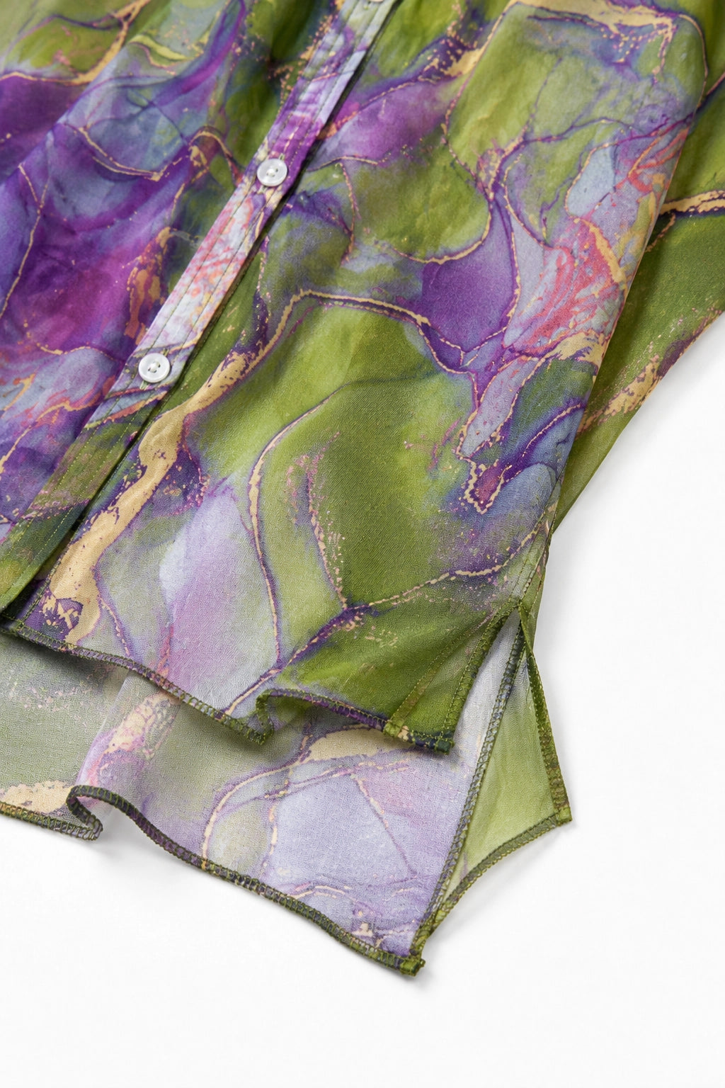 A close-up of a lightweight button-front garment with an abstract marbled print in green, purple, and lavender tones, with metallic gold veining and a white button visible.