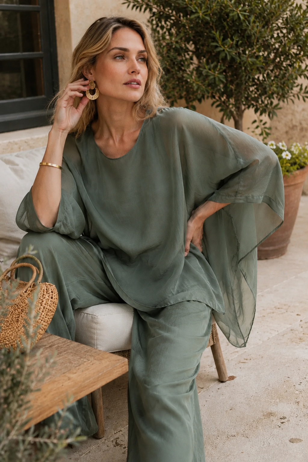 A woman seated outdoors wearing a flowing sage green two-piece outfit with a loose top and wide-leg pants. The setting includes a stone patio, potted plants, a woven bag, and a wooden bench.