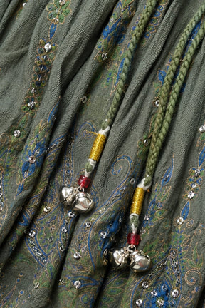 Close-up of a green patterned fabric with blue and gold paisley motifs, embellished with small silver rhinestones and two braided drawstring cords ending in silver bells.