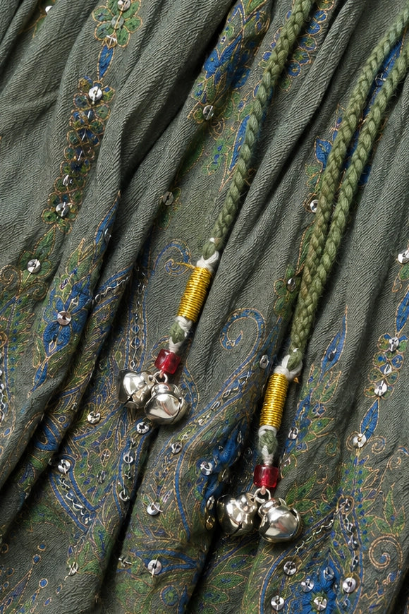 Close-up of a green patterned fabric with blue and gold paisley motifs, embellished with small silver rhinestones and two braided drawstring cords ending in silver bells.