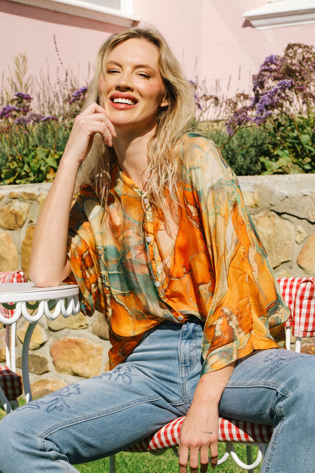 A woman sitting outdoors on a garden patio wearing a loose, semi-sheer orange and green abstract print blouse with blue jeans.