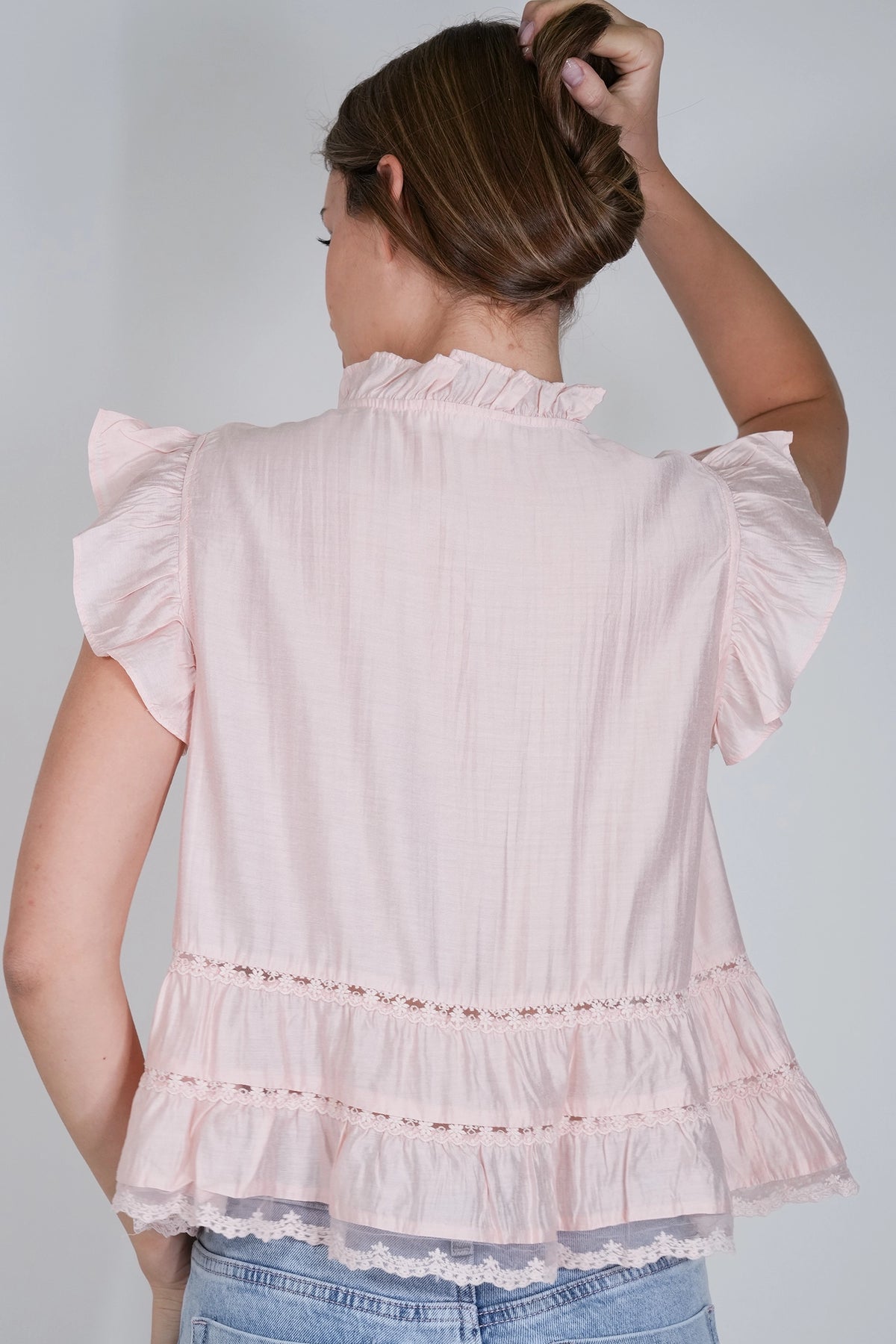 A woman shown from the back wearing a light pink ruffled blouse with short flutter sleeves and a tiered ruffle hem, paired with light blue jeans against a plain light gray background.