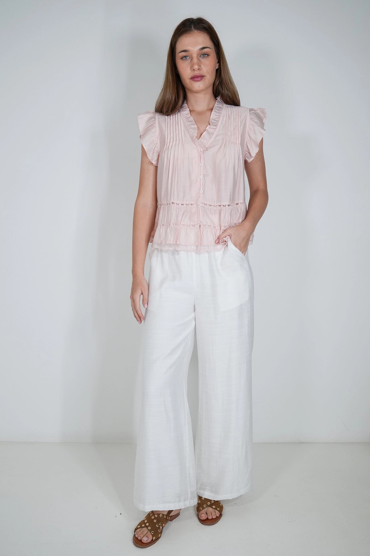 A woman modeling a light pink pleated ruffle-sleeve blouse paired with white wide-leg pants and brown sandals, standing against a plain white studio background.