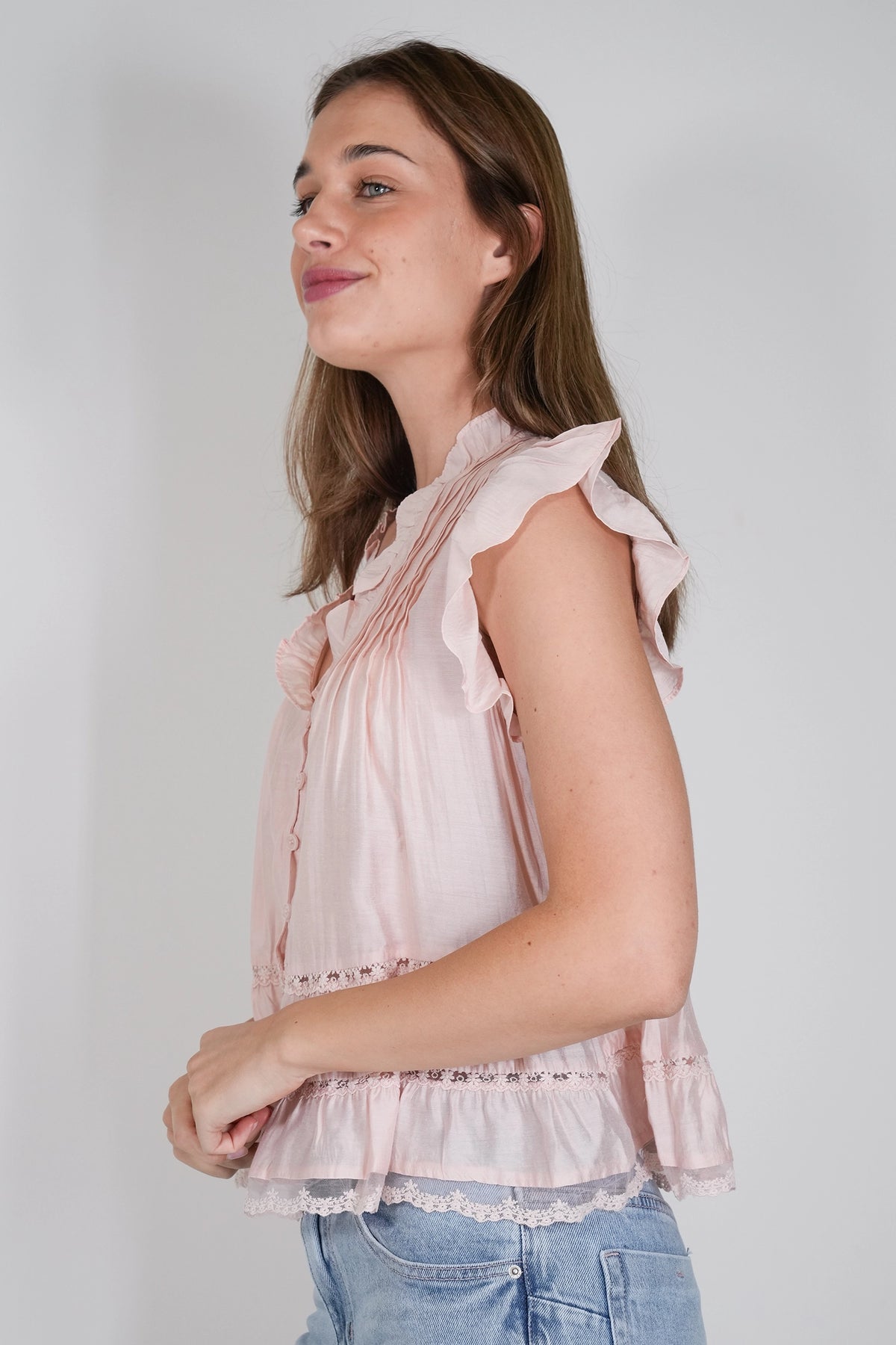 A woman wearing a light pink sleeveless blouse with ruffled shoulders and a gathered peplum hem, paired with light blue jeans, photographed against a plain light gray background.