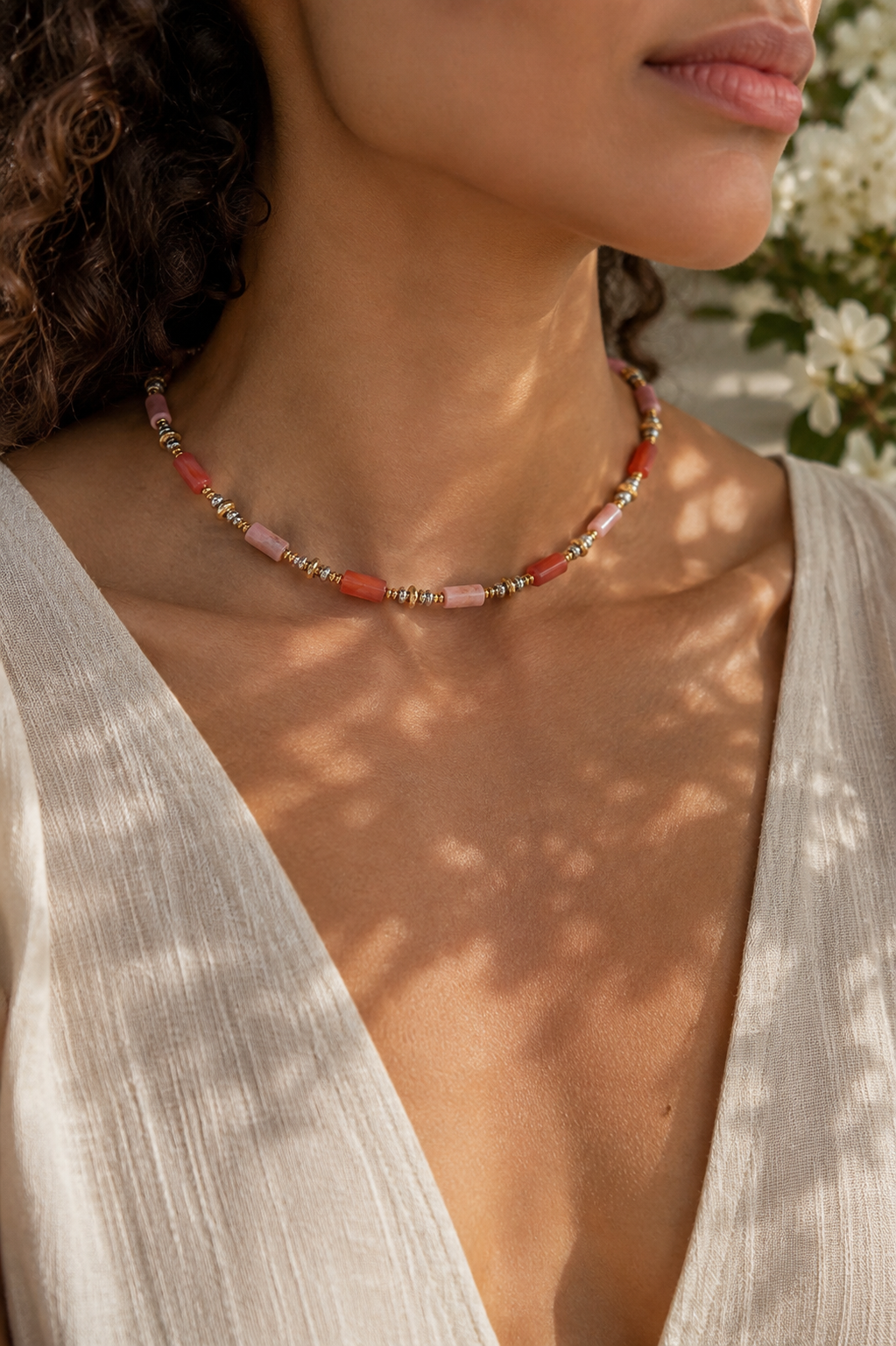 A close-up lifestyle shot of a woman wearing a delicate beaded necklace with pink, orange, and gold beads, styled with a cream-colored V-neck top.
