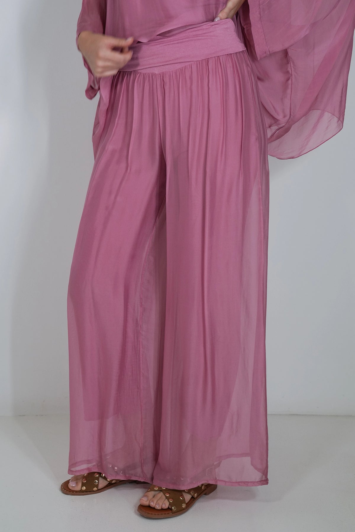 A person wearing loose, flowing mauve-pink wide-leg pants with a matching waist tie and a sheer long-sleeve top, paired with brown sandals, against a plain white studio background.