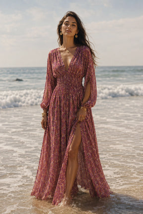 A woman wearing a long, dusty rose floral maxi dress with a thigh-high slit stands on a sandy beach with the ocean waves in the background.