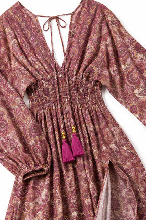 A close-up of a long-sleeve bohemian-style dress with a smocked waist, deep V-neckline with tie details, and bright magenta tassel accents, shown on a white background.