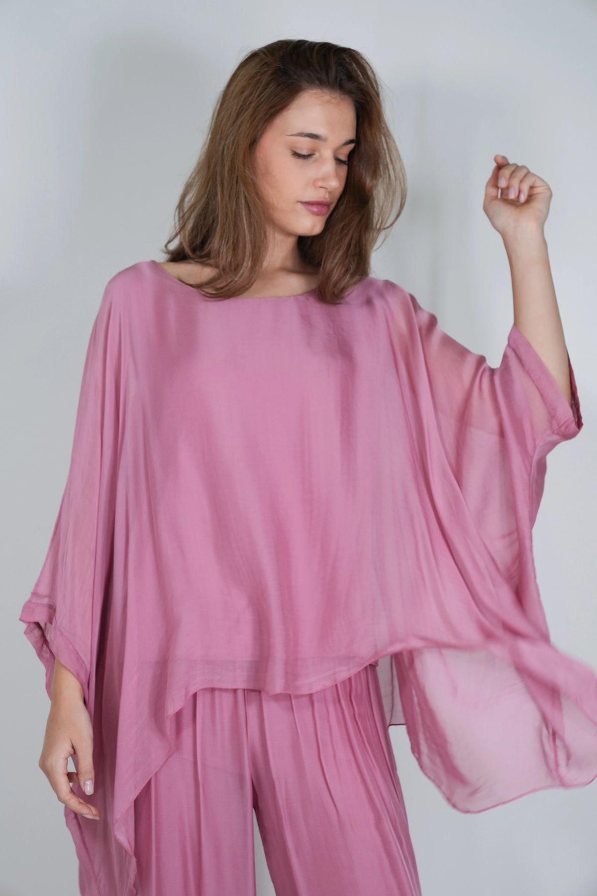 A woman wearing a flowing dusty pink two-piece outfit with a loose, draped top and matching pants, photographed against a plain light gray studio background.