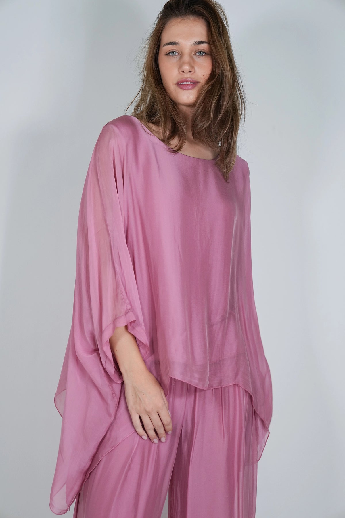 A woman modeling a matching dusty pink two-piece outfit consisting of a loose, semi-sheer long-sleeve top and coordinating wide-leg trousers, set against a plain light gray studio background.
