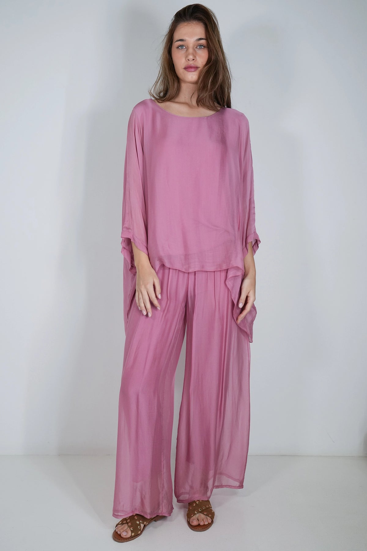 A woman modeling a dusty pink two-piece outfit consisting of a loose, long-sleeve top and matching wide-leg pants, standing against a plain white studio background.