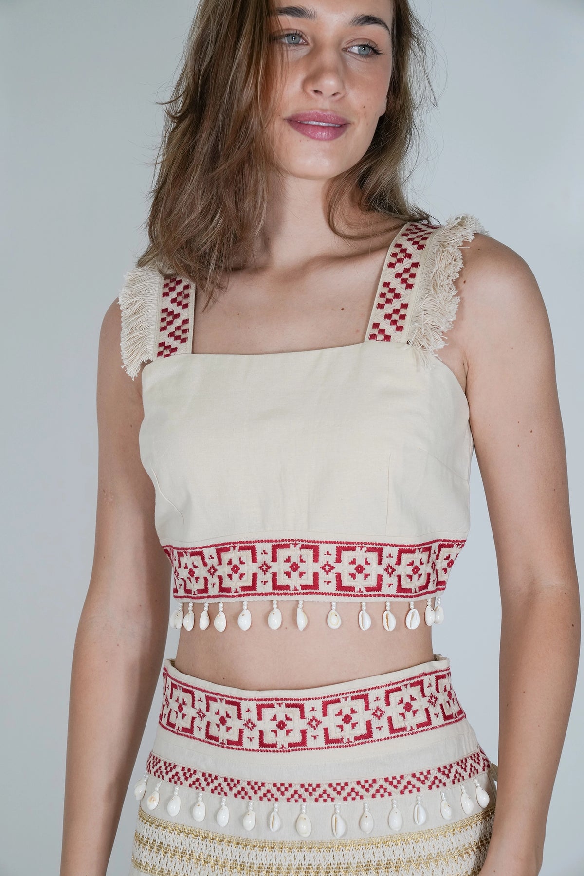 A woman wearing a cream-colored crop top with red geometric embroidery, fringed straps, and tassel trim, paired with a matching patterned skirt, against a plain light gray background.