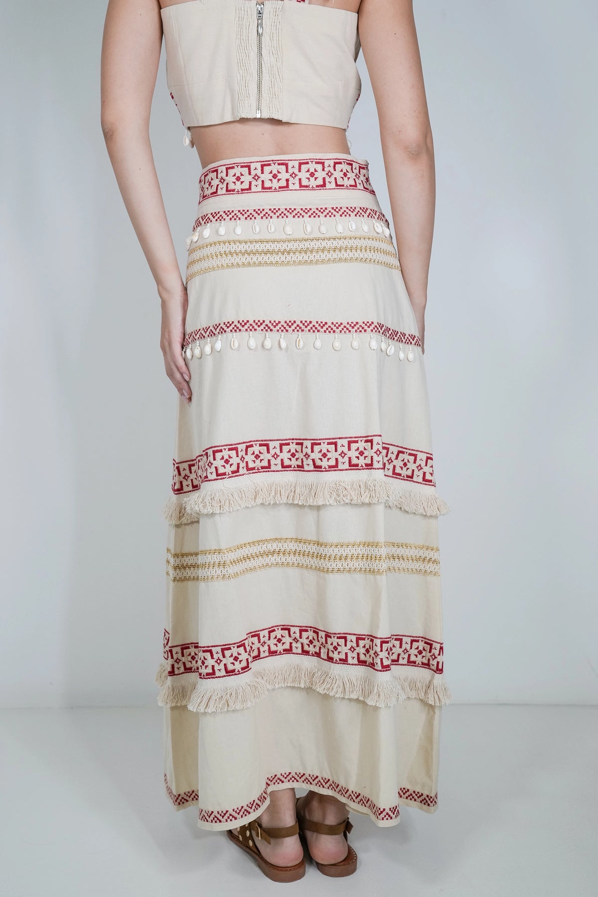 A model is shown from the waist down wearing a cream-colored bohemian maxi skirt with red and beige geometric patterns, fringe details, and a matching cream strapless crop top against a light gray studio background.
