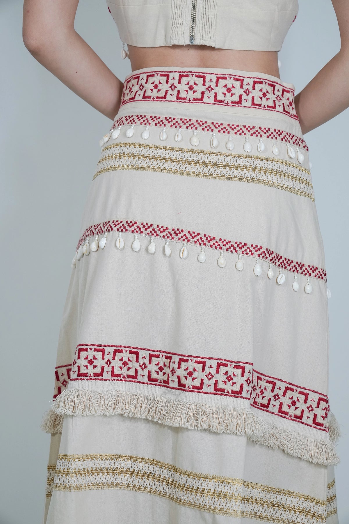 A close-up of a cream-colored bohemian maxi skirt with red and gold geometric embroidery, shell bead embellishments, and fringe details, worn by a model.