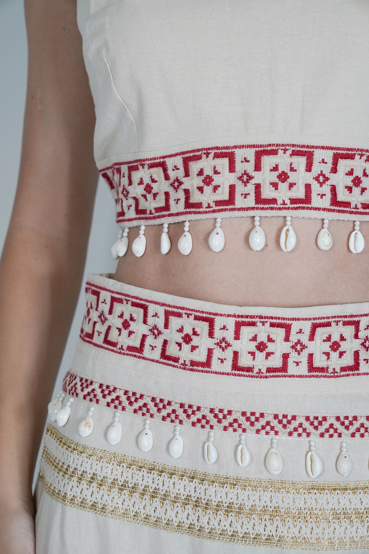 Close-up of a person wearing a cream-colored embroidered garment with red geometric patterns and white cowrie shell fringe.