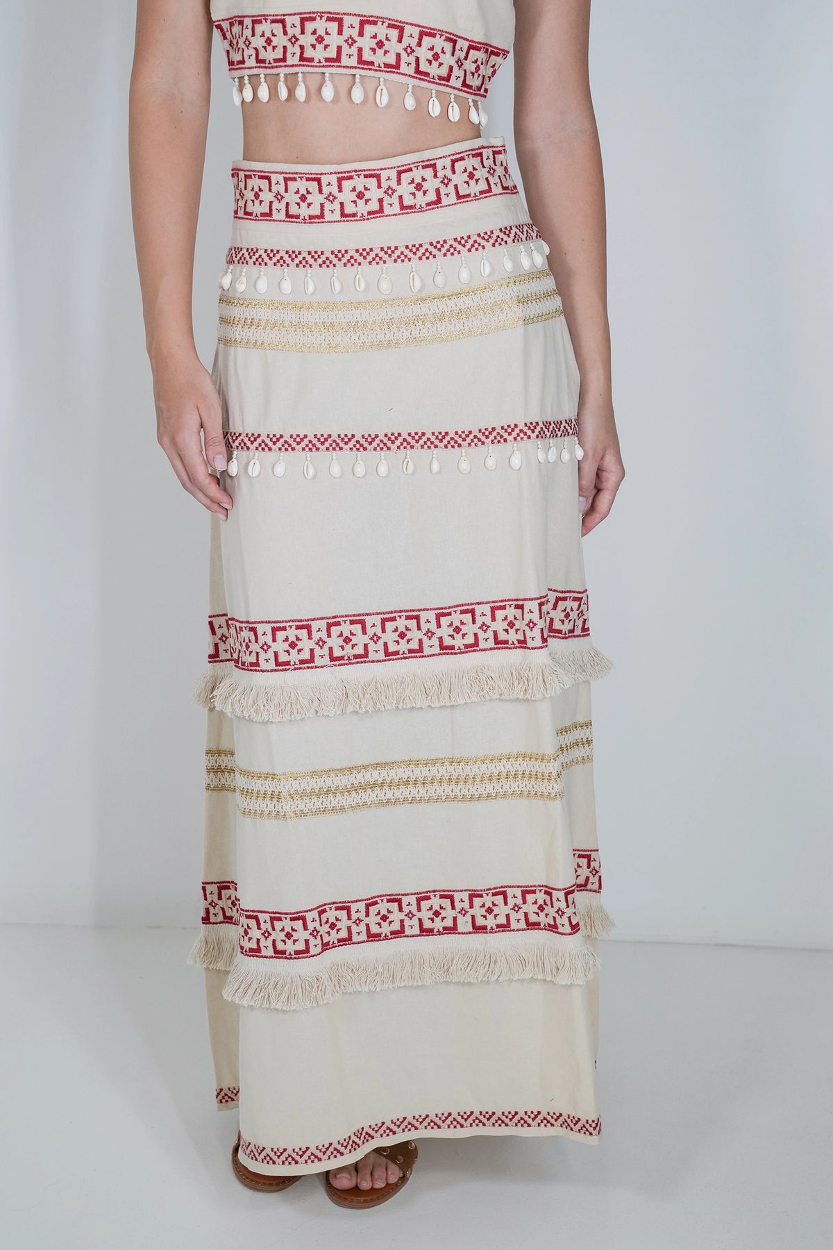 A model wearing a cream-colored bohemian maxi skirt with red and beige geometric patterns, tassels, and fringe, paired with a matching crop top against a plain white studio background.