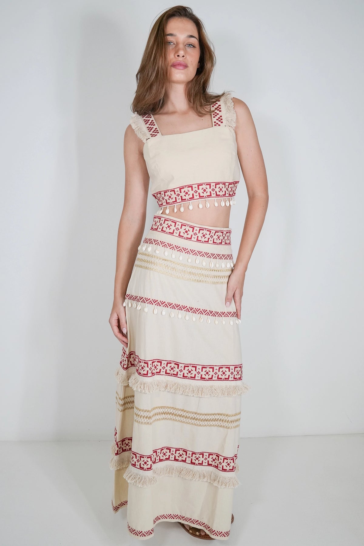 A woman modeling a sleeveless cream maxi dress with red and beige bohemian-style embroidered horizontal bands and tassel trim, set against a plain light gray studio background.
