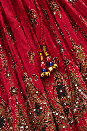 A close-up of a rich red fabric with intricate gold, brown, and black paisley embroidery, adorned with small mirror work and decorative tassels with red, blue, and gold beads.