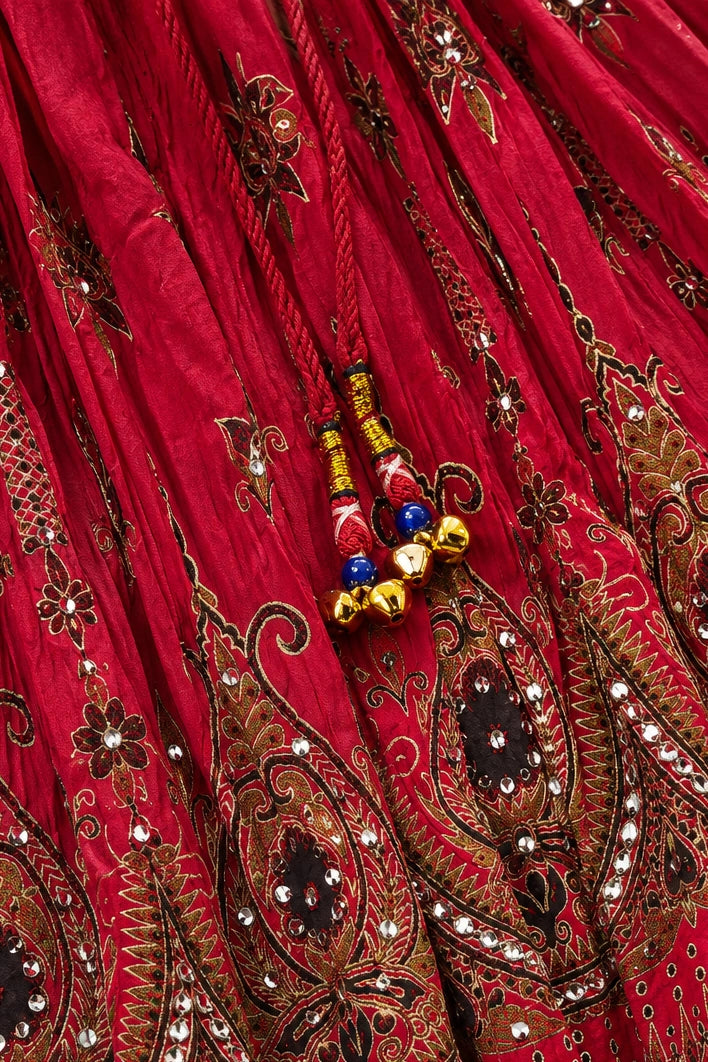 A close-up of a rich red fabric with intricate gold, brown, and black paisley embroidery, adorned with small mirror work and decorative tassels with red, blue, and gold beads.