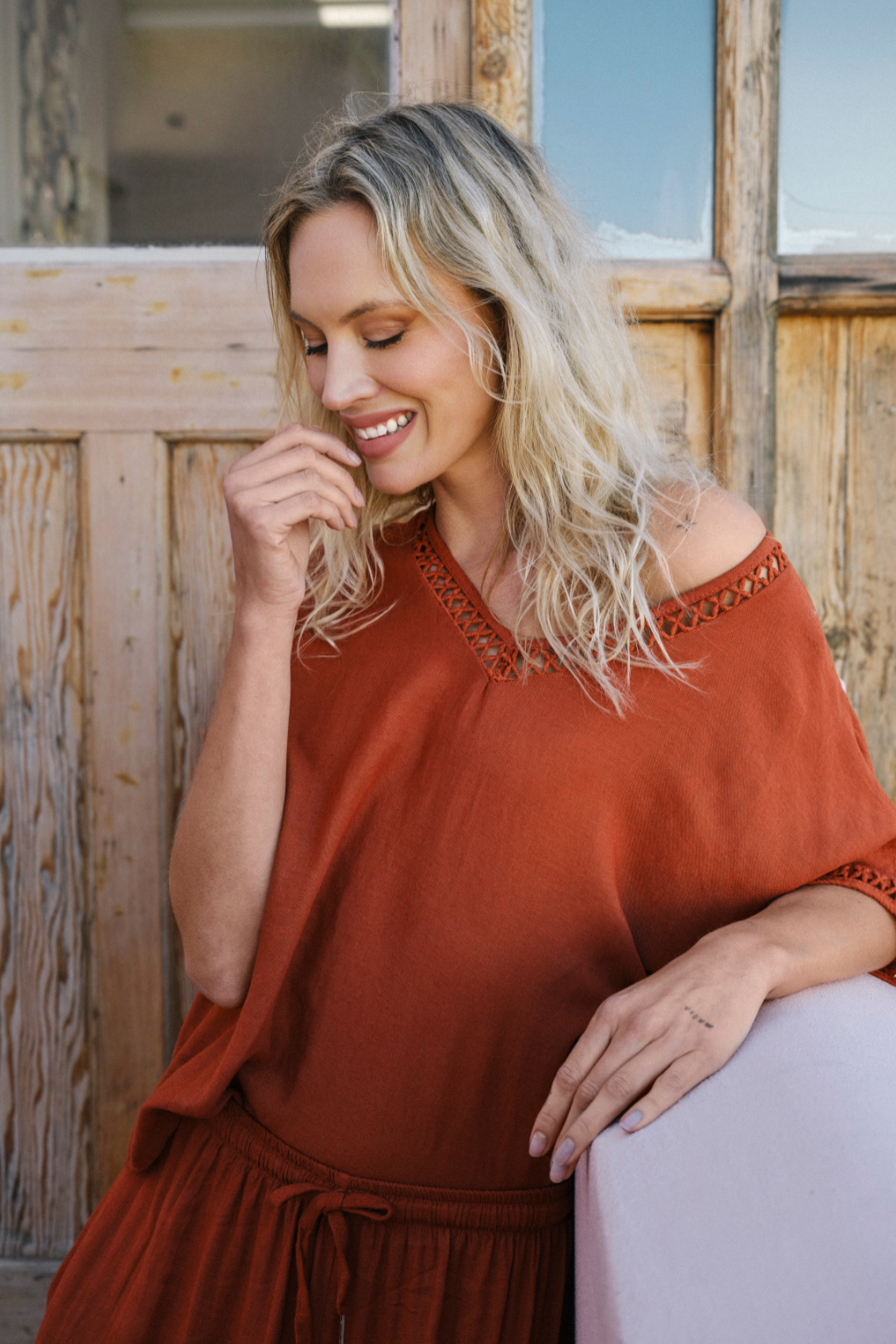 A woman with blonde hair wearing a rust-orange off-the-shoulder top and matching skirt, leaning against a white chair in front of a rustic wooden door.