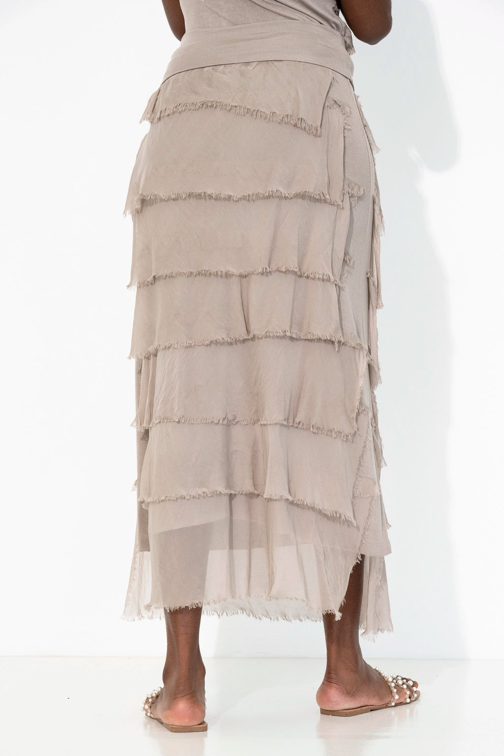 A model from the back wearing a tiered, ruffled midi skirt in a light taupe-beige color, paired with flat sandals against a white studio background.