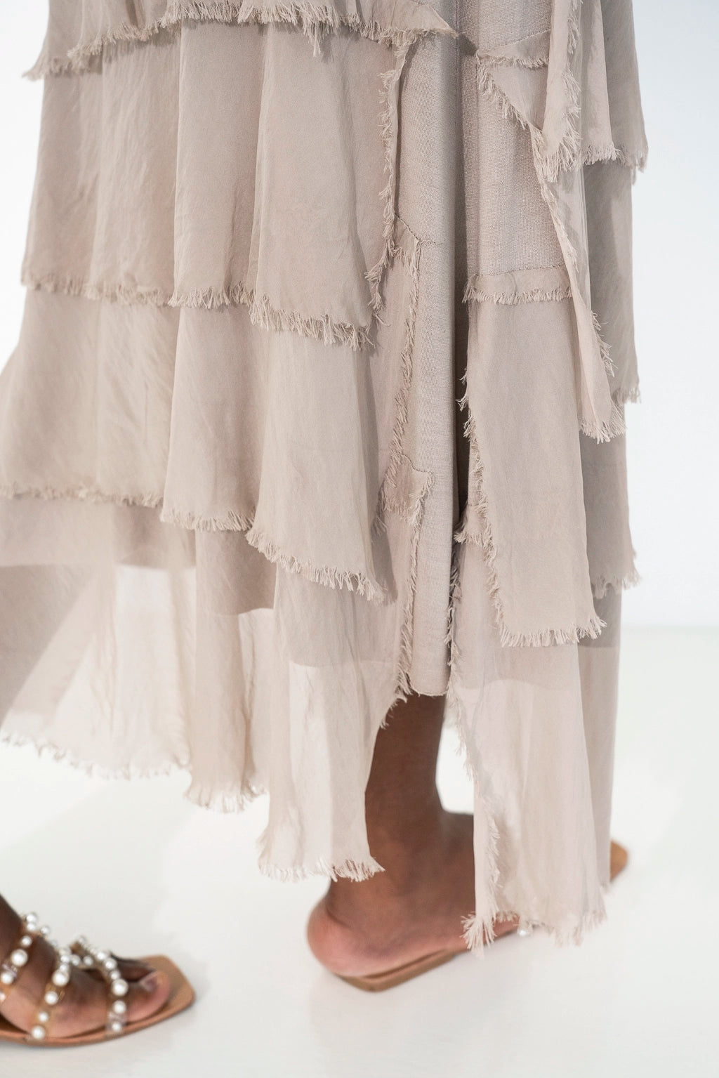 A close-up of a person wearing a long, tiered, frayed-edge maxi skirt in a light taupe-beige color, paired with brown slide sandals with pearl embellishments.
