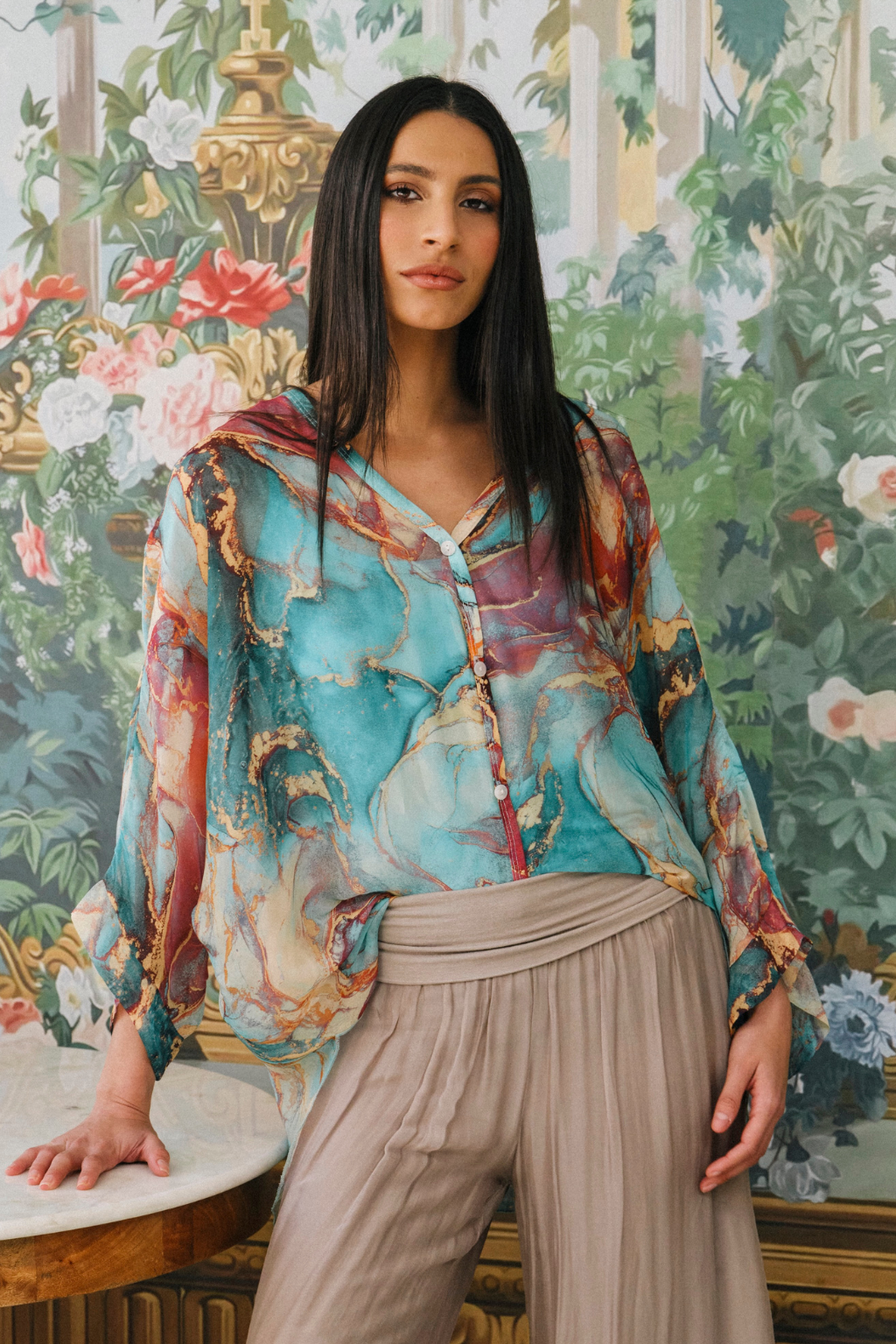 A woman wearing a long-sleeve V-neck blouse with a colorful abstract print in turquoise, red, purple, and gold, paired with beige wide-leg trousers, posed in front of a detailed classical floral wallpaper background.