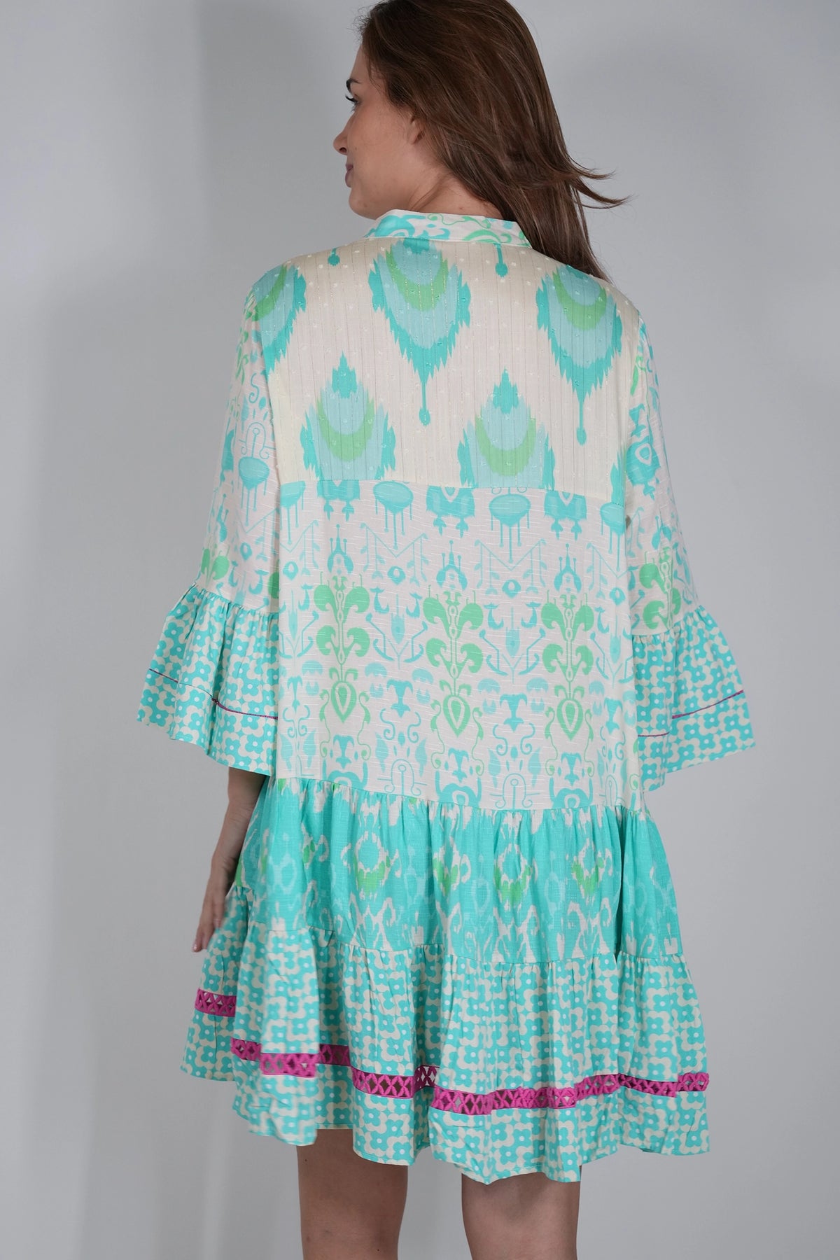 A woman shown from the back wearing a tiered, short dress with a white base and a vibrant turquoise, green, and pink ikat pattern.