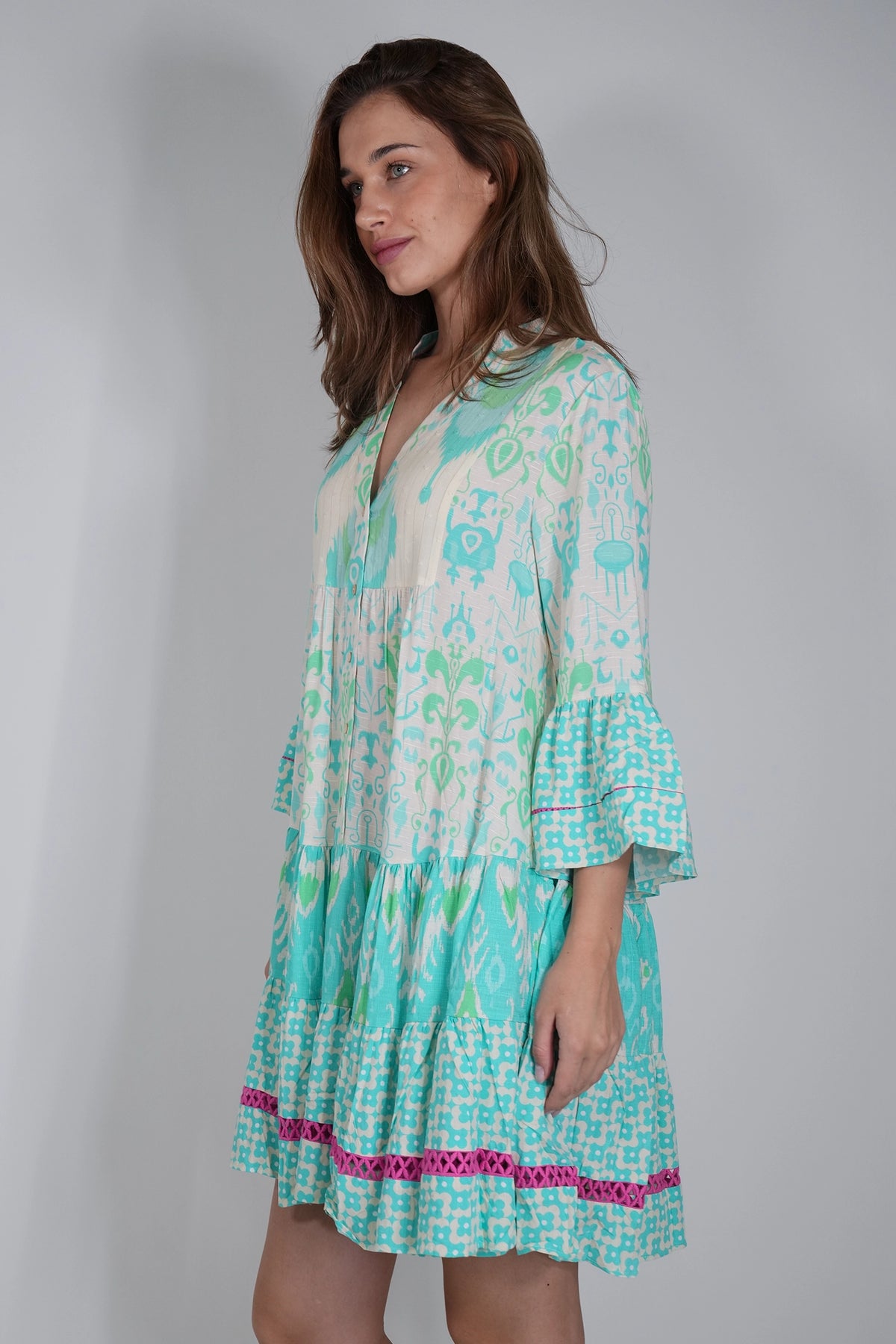 A woman modeling a tiered short dress with a white base and a turquoise and green abstract pattern, featuring a pink leopard-print ruffle trim at the hem, set against a plain light gray studio background.