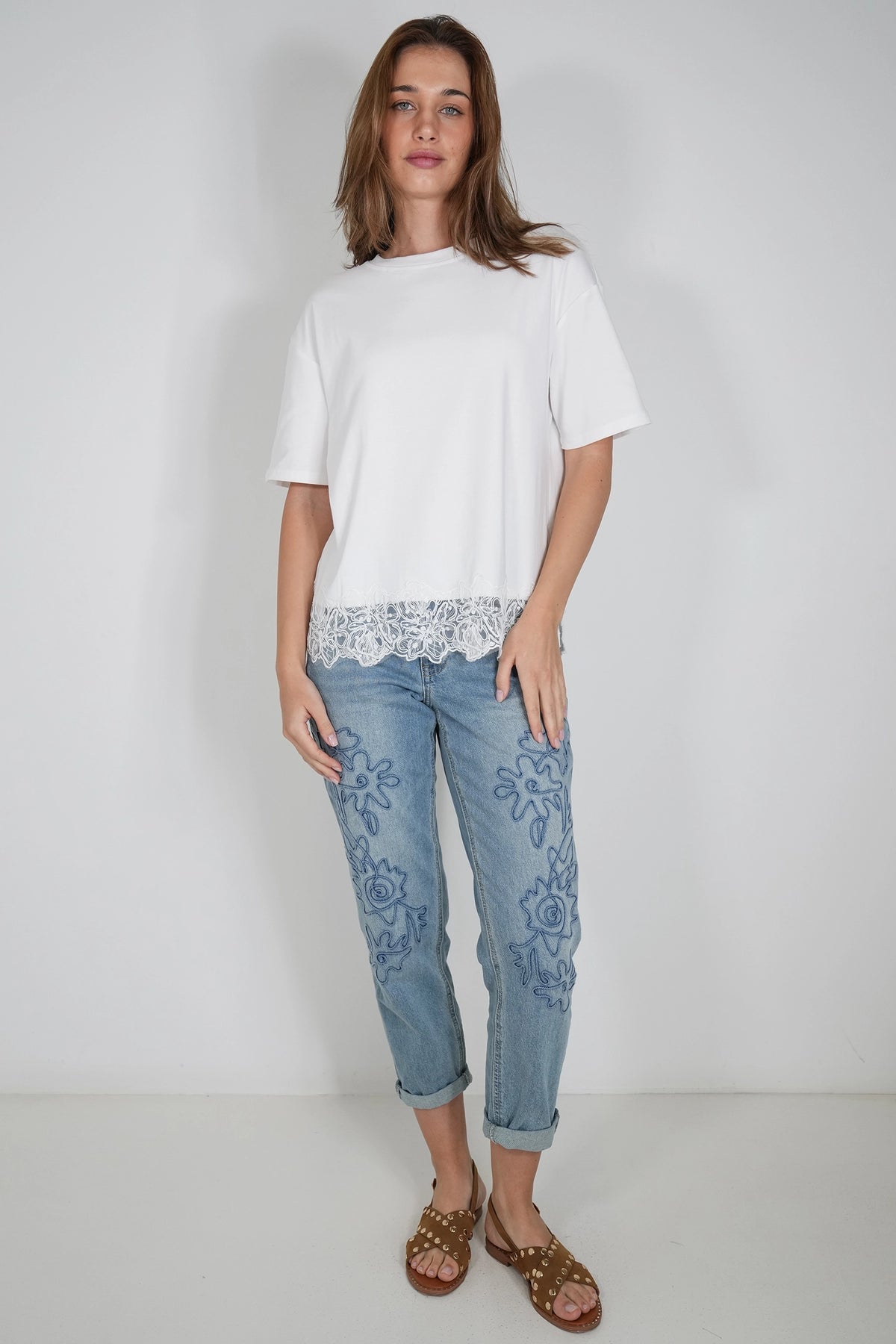 A woman modeling a white short-sleeve t-shirt with a lace hem, paired with light blue floral embroidered jeans and brown studded sandals, against a plain light gray studio background.