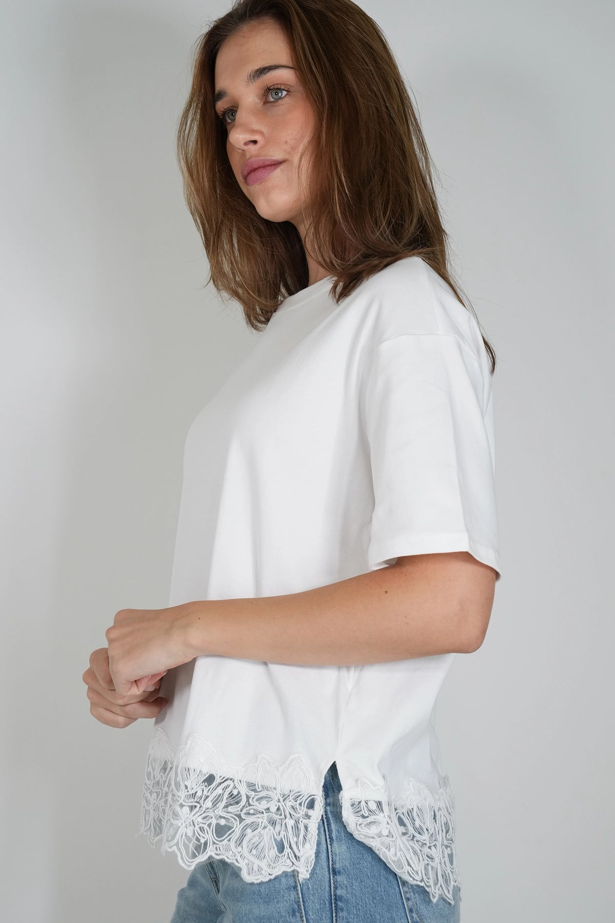 A woman wearing a white short-sleeve top with a lace hem detail, paired with light blue jeans, photographed against a plain light gray studio background.