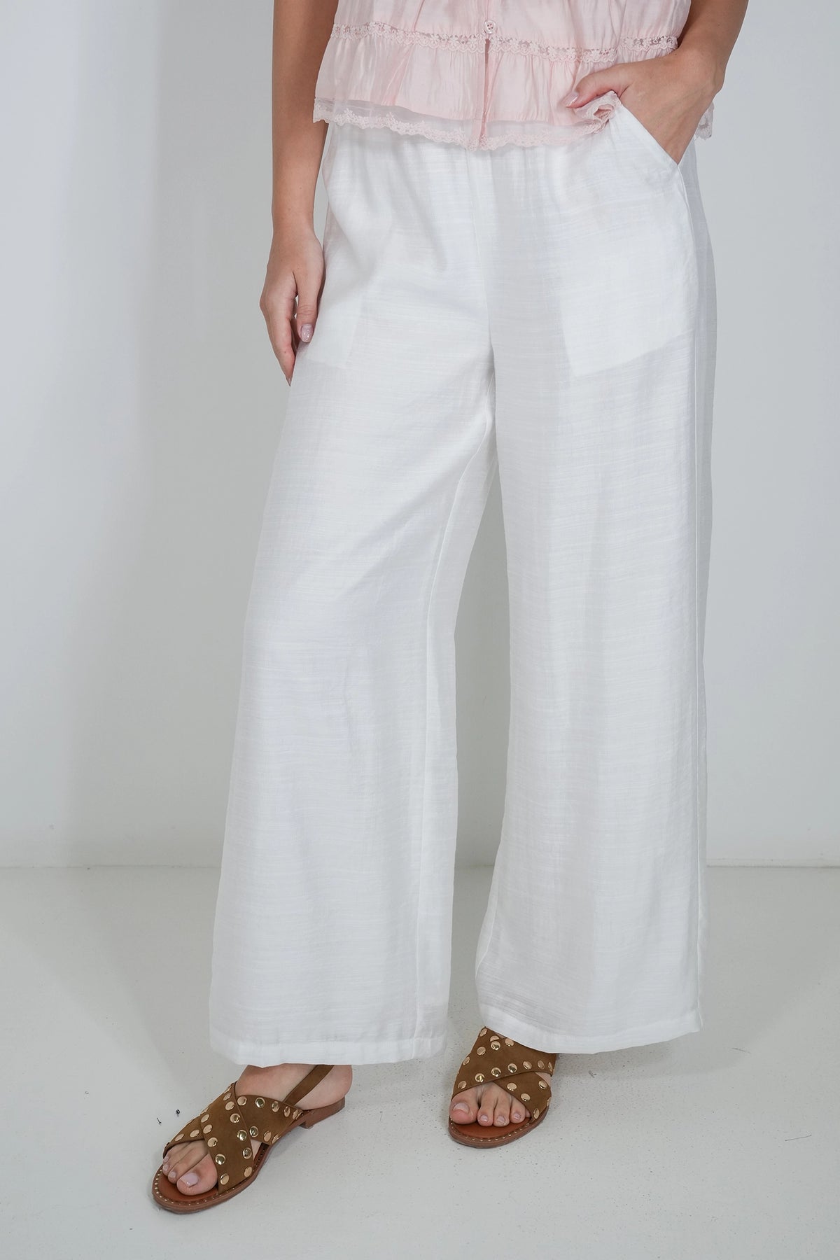 White wide-leg linen pants worn by a model, styled with a light pink ruffled top and brown studded sandals against a white studio background.