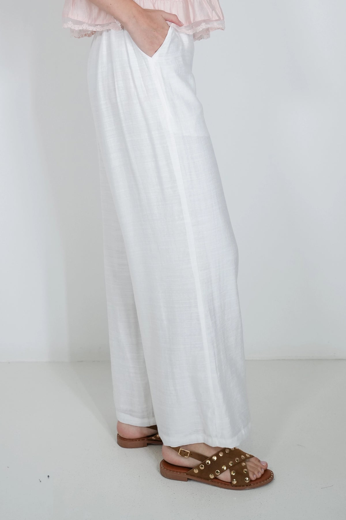 White wide-leg linen pants worn by a model, paired with brown studded sandals and a light pink ruffled top, shown against a plain white studio background.