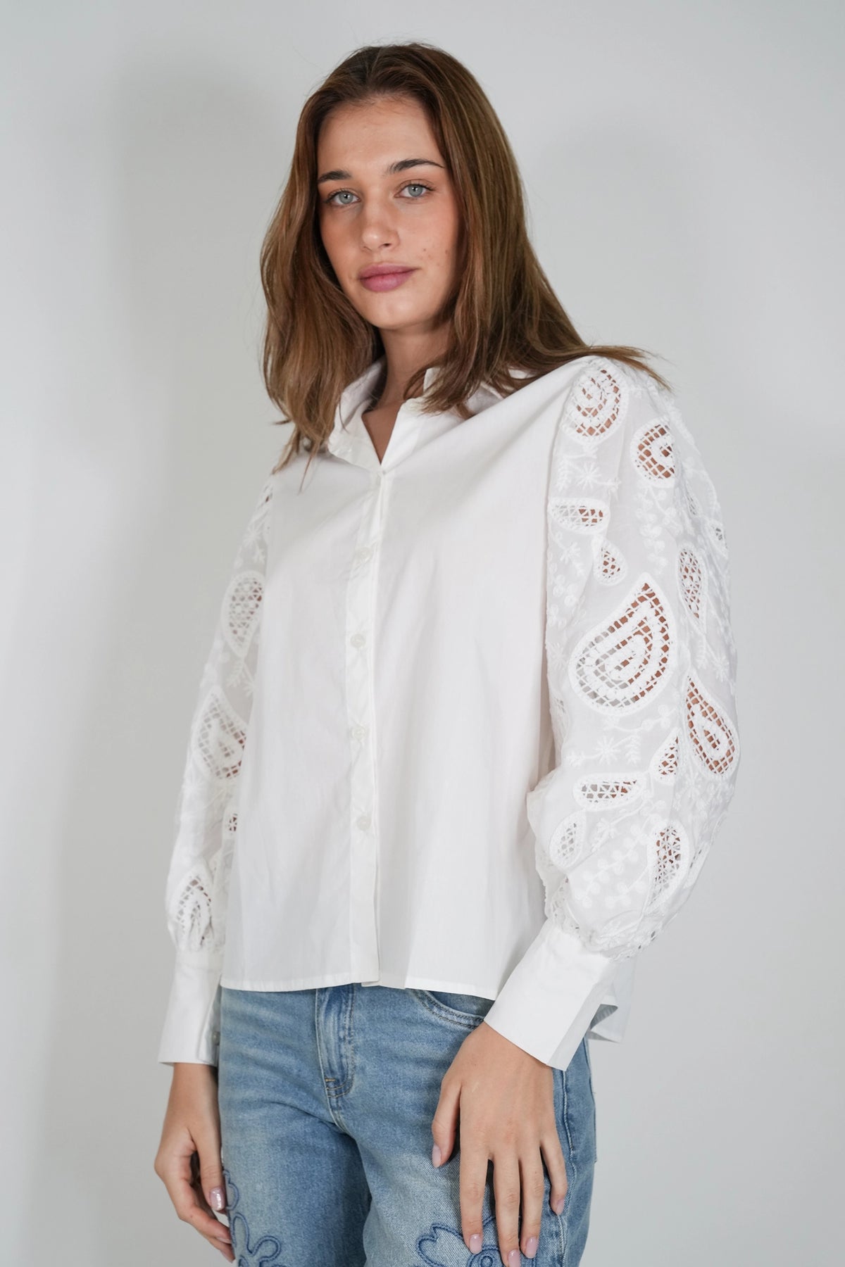 A woman wearing a white long-sleeve button-up blouse with intricate eyelet lace detailing on the sleeves, paired with blue jeans, photographed against a neutral light gray background.