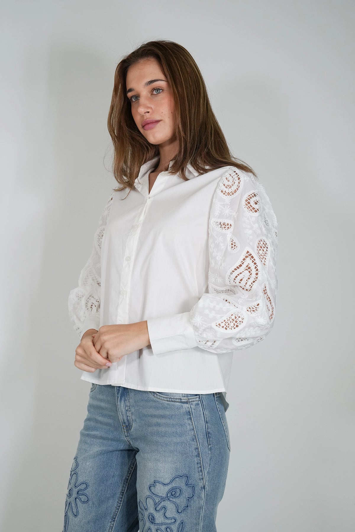 A woman wearing a white long-sleeve button-up blouse with intricate floral eyelet lace detailing on the sleeves, paired with light blue jeans featuring embroidered floral patterns, set against a plain light gray studio background.