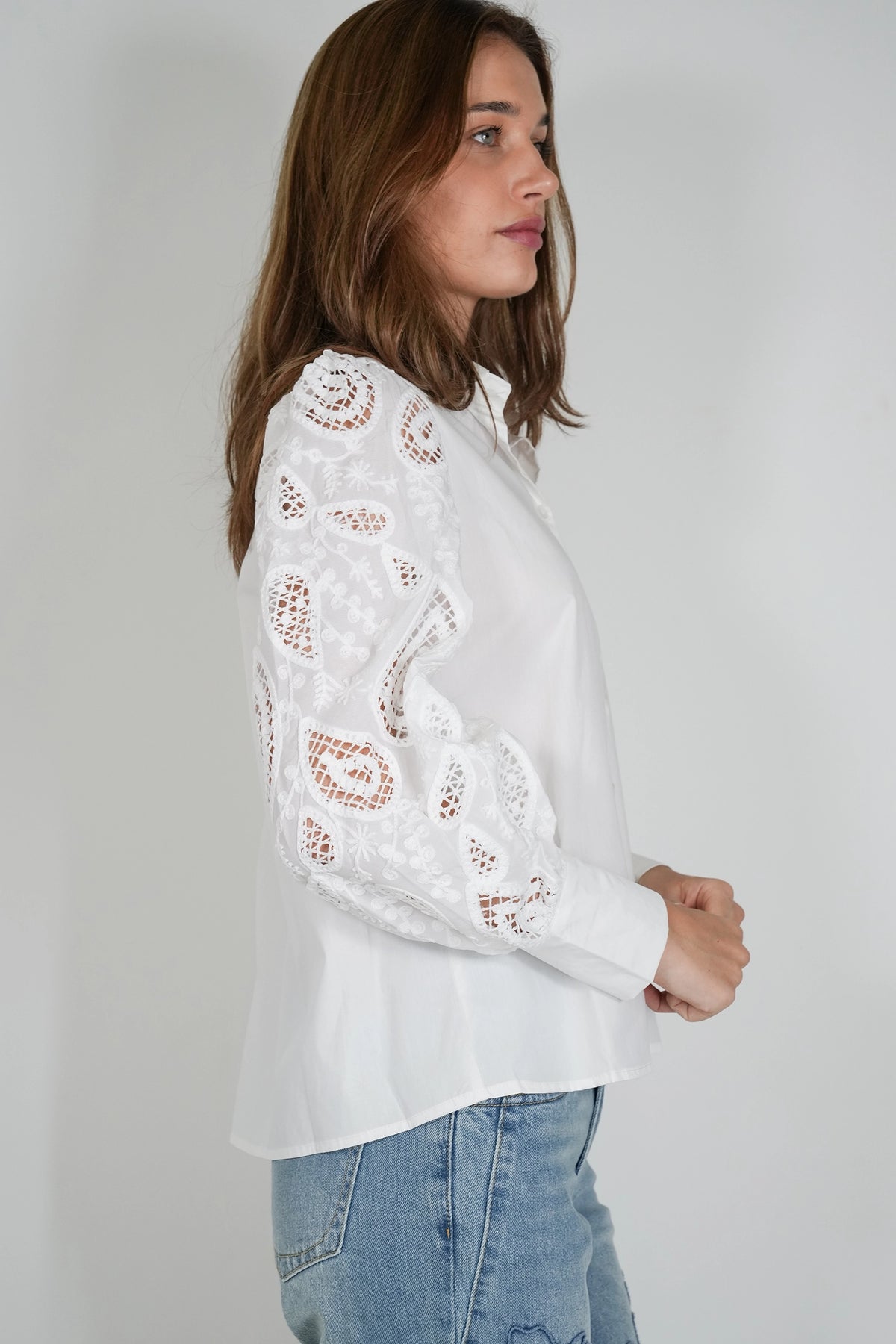 A woman wearing a white long-sleeve blouse with intricate lace and eyelet embroidery on the sleeves, paired with light blue jeans, shown from a side profile against a plain light gray background.