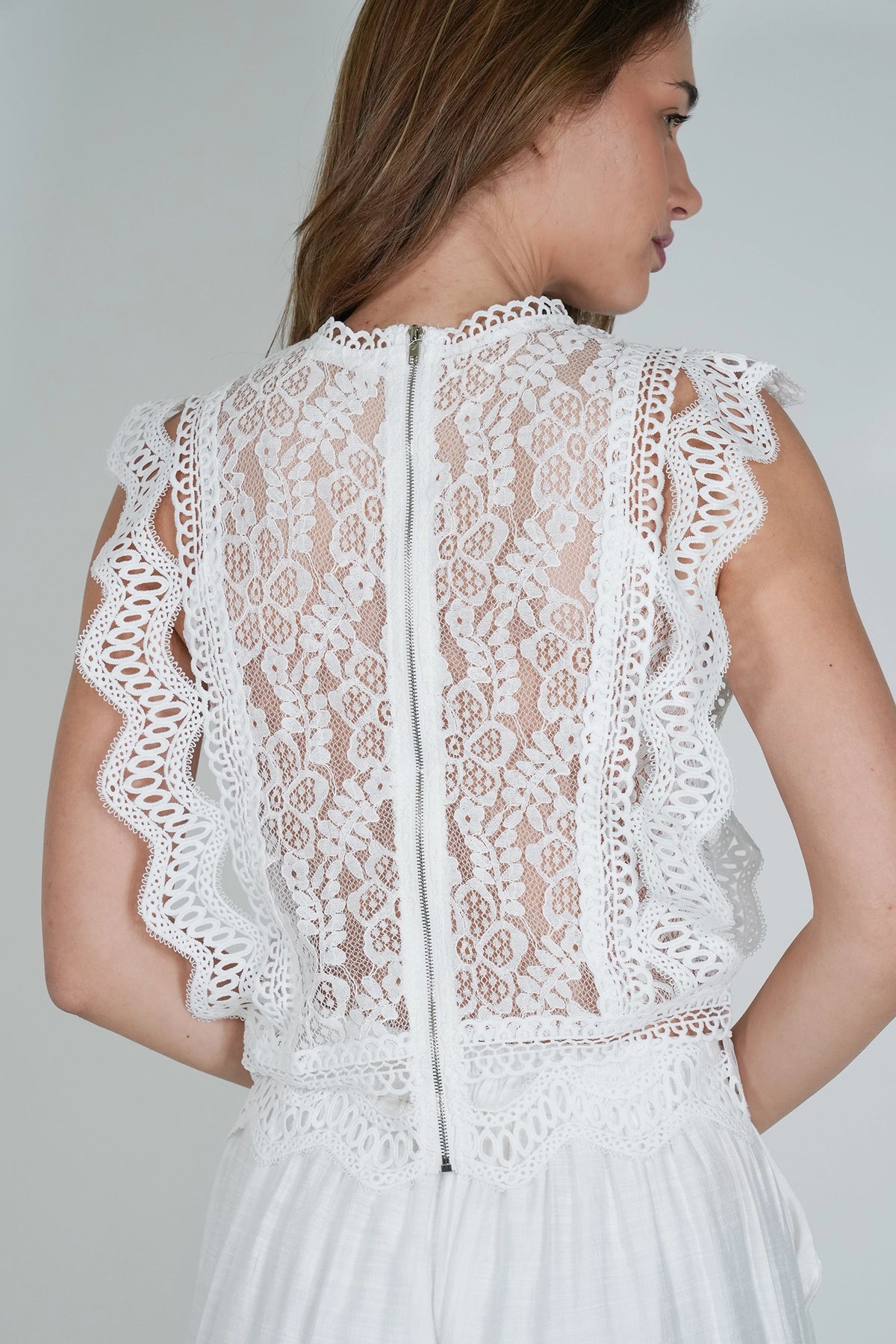 A woman shown from the back wearing a white sleeveless lace top with a high neckline, scalloped lace trim, and a central zipper, against a light gray studio background.
