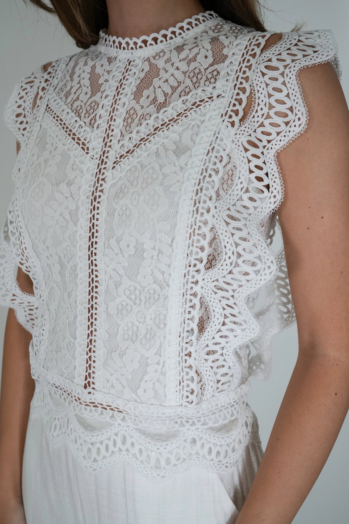White sleeveless lace top with a high mock neck, scalloped edges, and intricate crochet detailing, shown on a model.