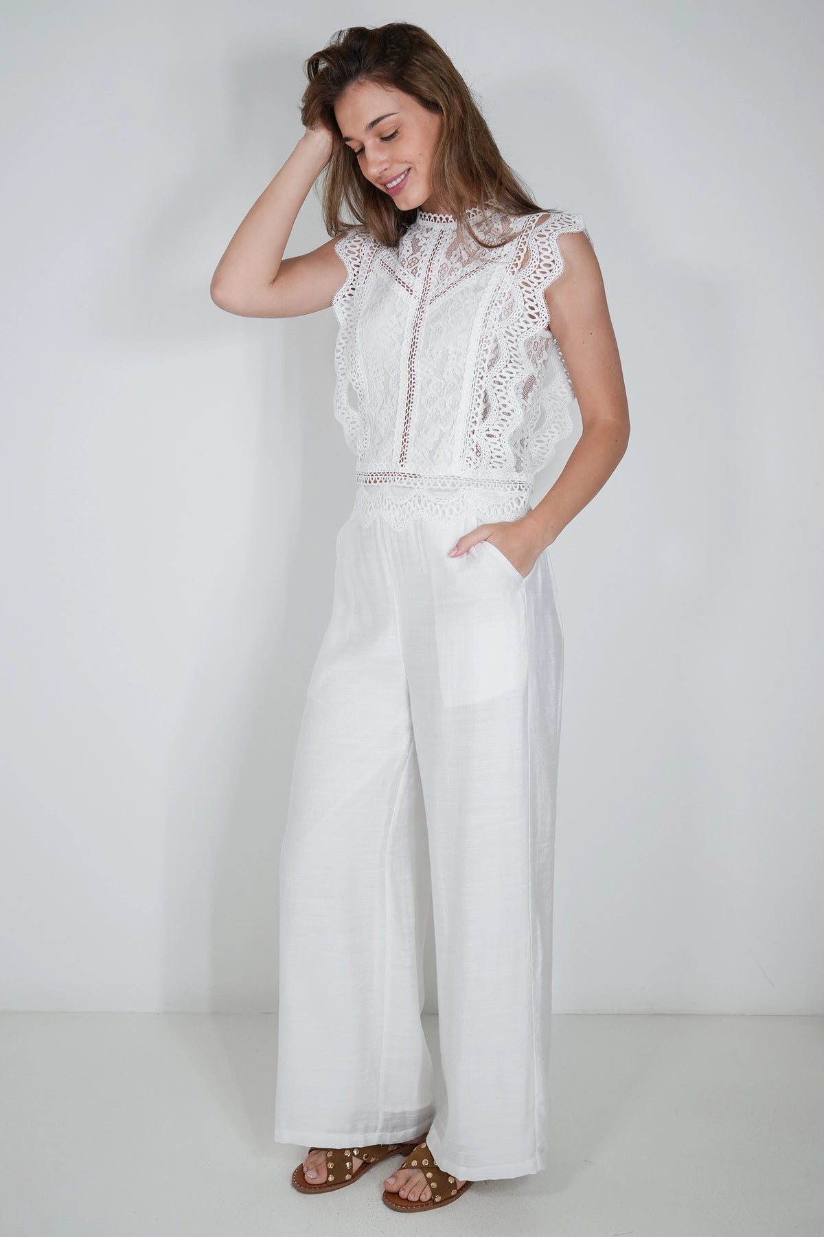 A woman modeling a white sleeveless lace-trimmed top and matching wide-leg pants set, paired with brown strappy sandals, against a plain white studio background.