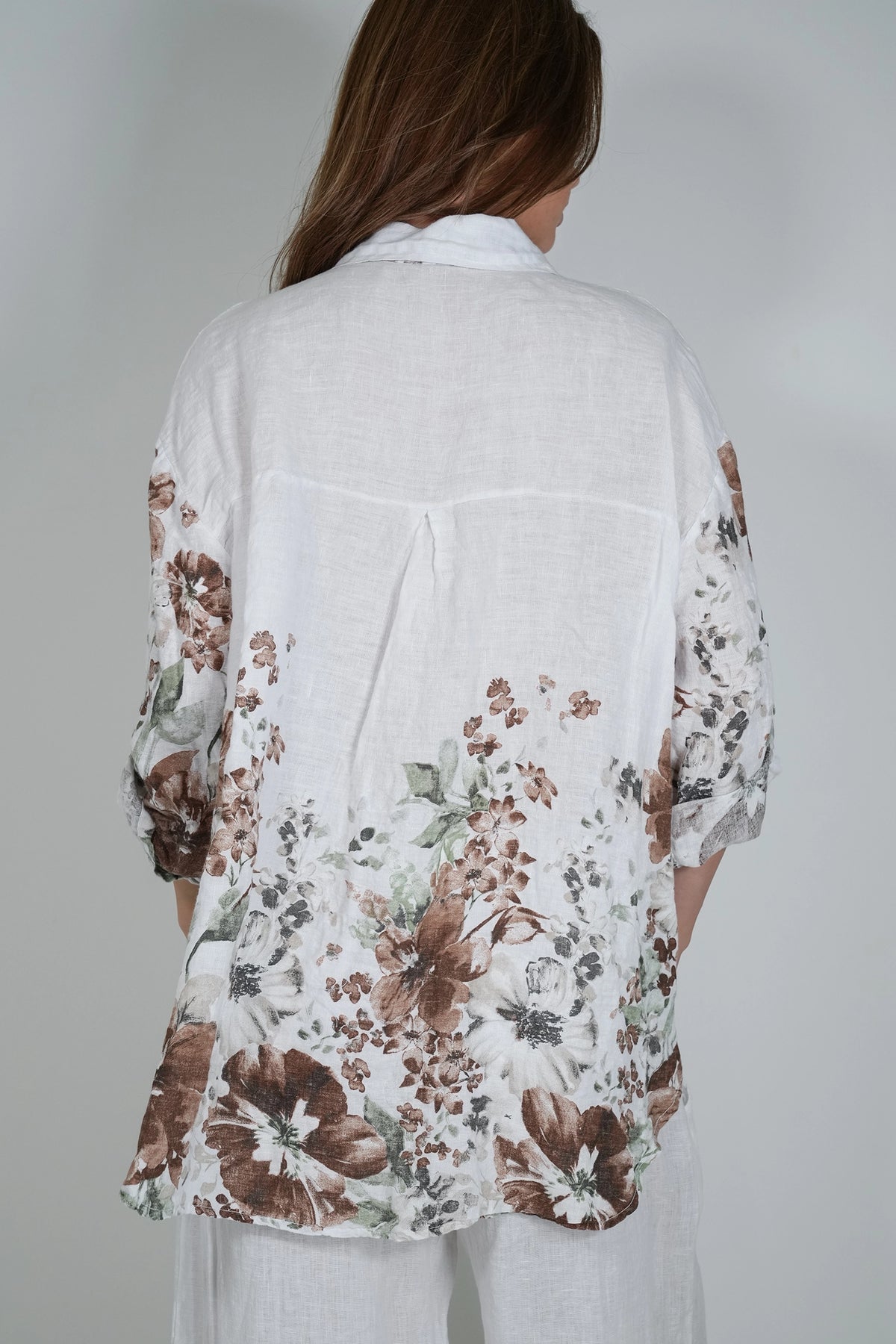 A woman viewed from behind wearing a loose white linen shirt with a brown and green floral print on the sleeves and lower back, paired with white pants, against a light gray studio background.