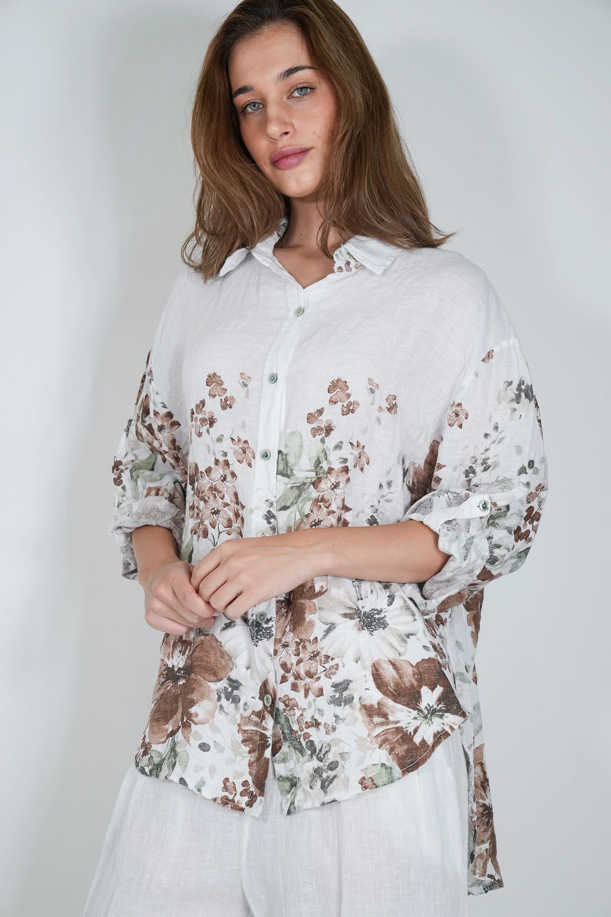 A woman wearing a white button-up shirt with a brown and green floral print, paired with white trousers, photographed against a light gray studio background.