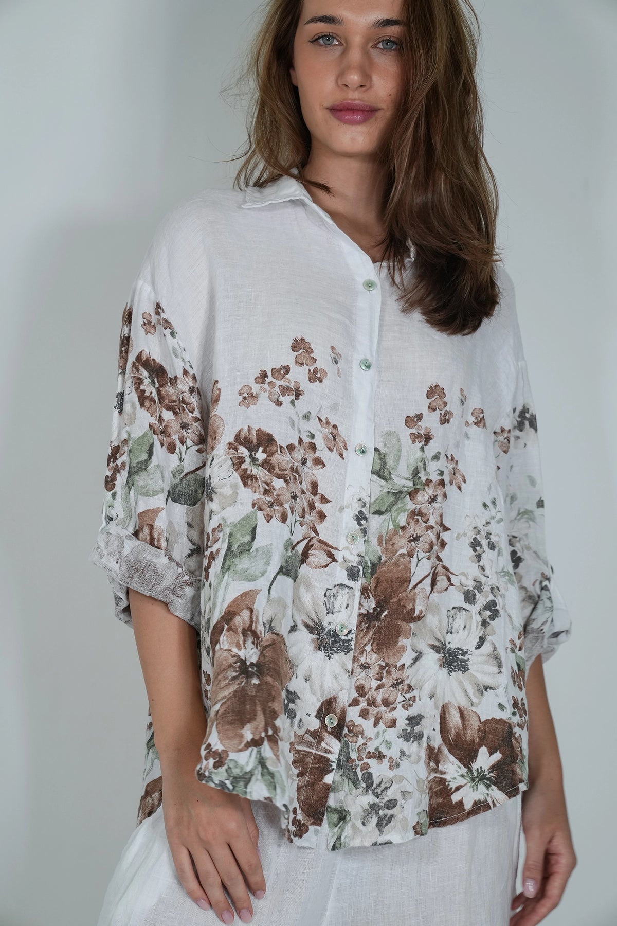 A woman wearing a loose white button-up blouse with a brown and green floral print, paired with white trousers, photographed against a light gray studio background.